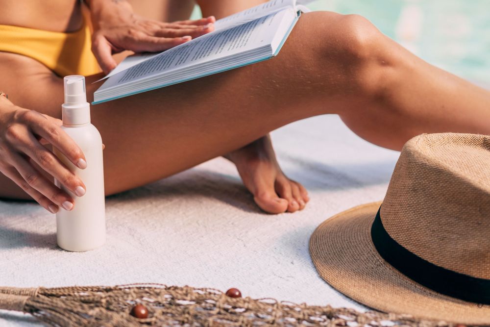 Frau sitzt am Strand mit Sonnencreme und Hut, liest ein Buch