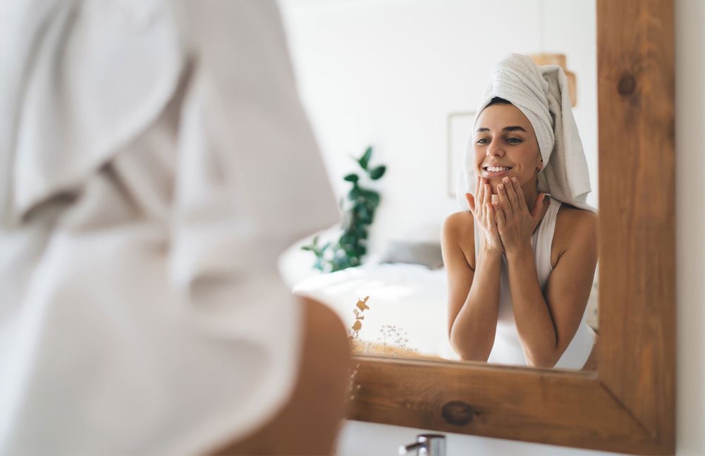Junge Frau im Handtuch trägt Gesichtspflege vor dem Spiegel auf
