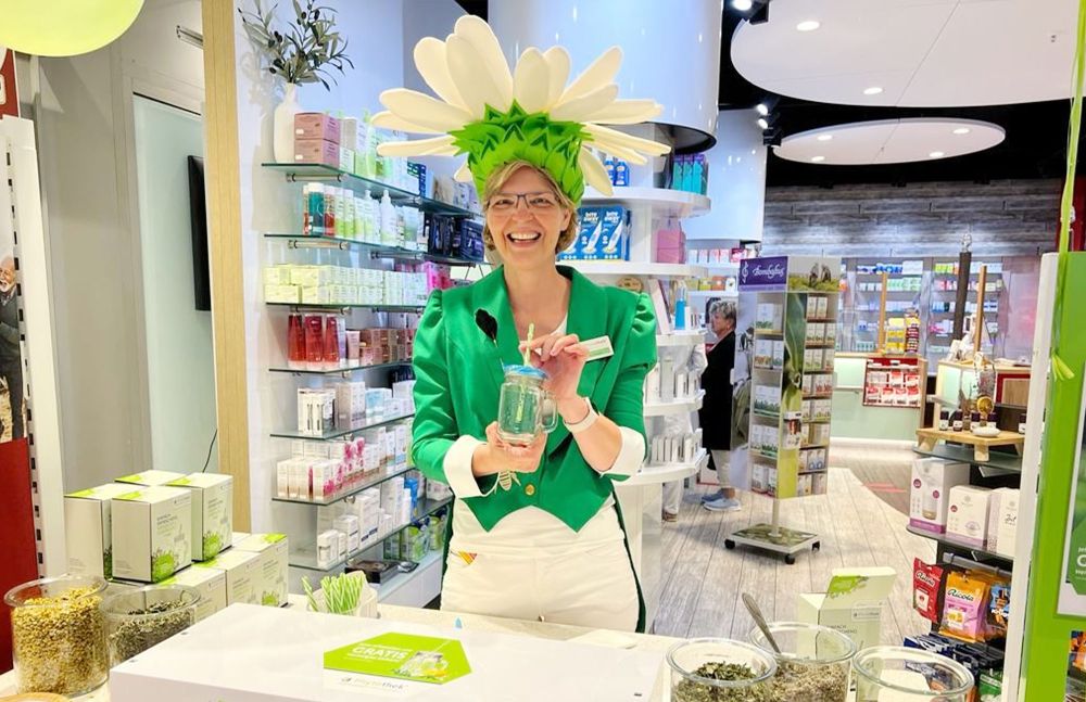Apothekerin mit Blütenhut lächelt am Aktionstisch in der Apotheke