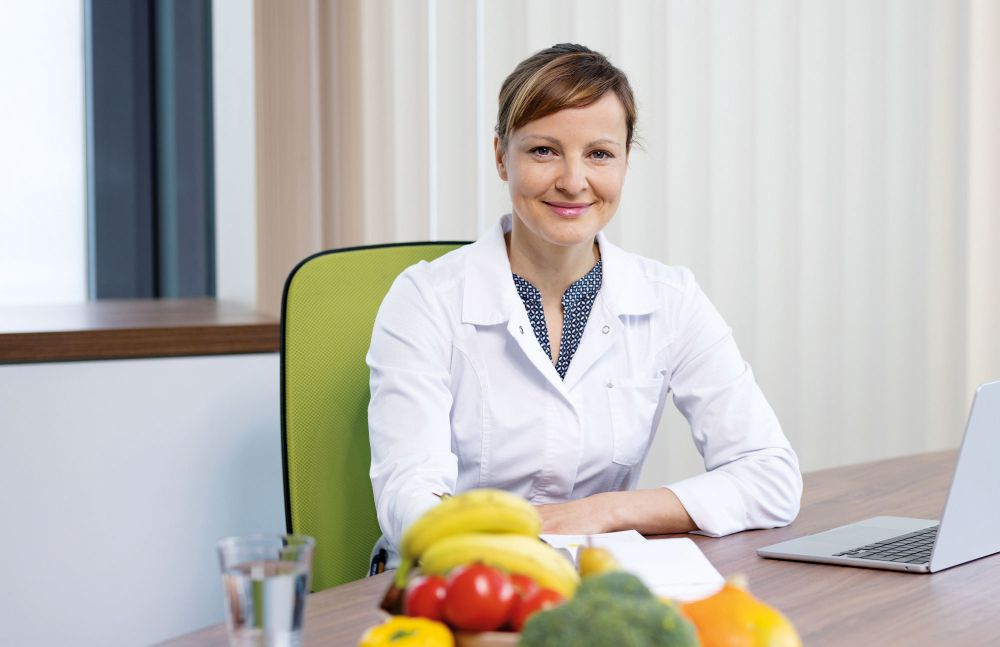 Ernährungsberaterin im weißen Kittel sitzt vor frischem Obst und Gemüse