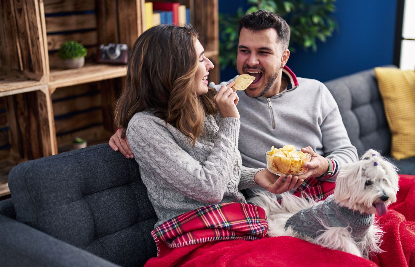Zwei Personen sitzen auf dem Sofa, essen Chips, kleiner Hund liegt daneben.