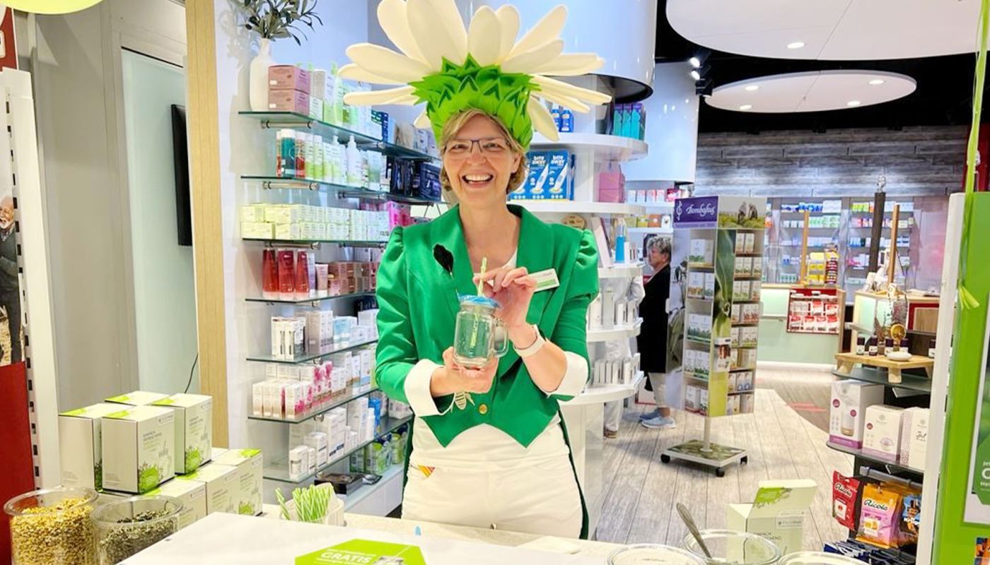 Apothekerin mit Blütenhut lächelt am Aktionstisch in der Apotheke