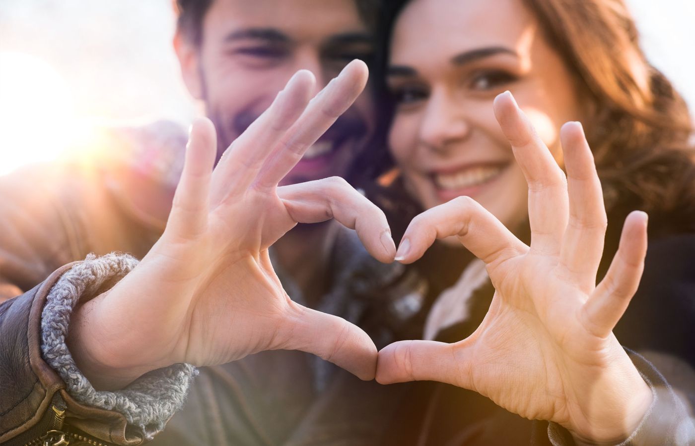 Zwei Personen halten ihre Hände zu einem Herz, Gesichter im Hintergrund unscharf.
