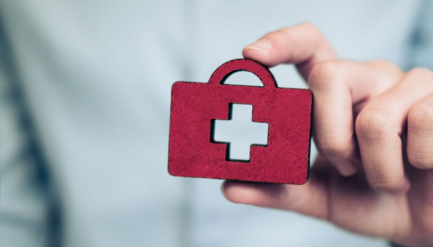 Hand hält rote Symbolfigur in Form einer Hausapotheke mit weißem Kreuz