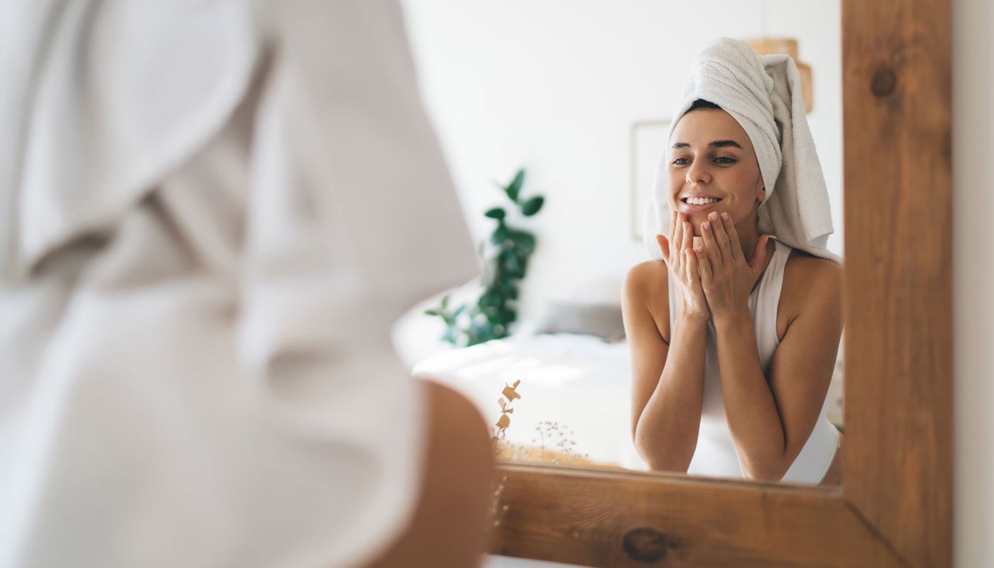 Junge Frau im Handtuch trägt Gesichtspflege vor dem Spiegel auf