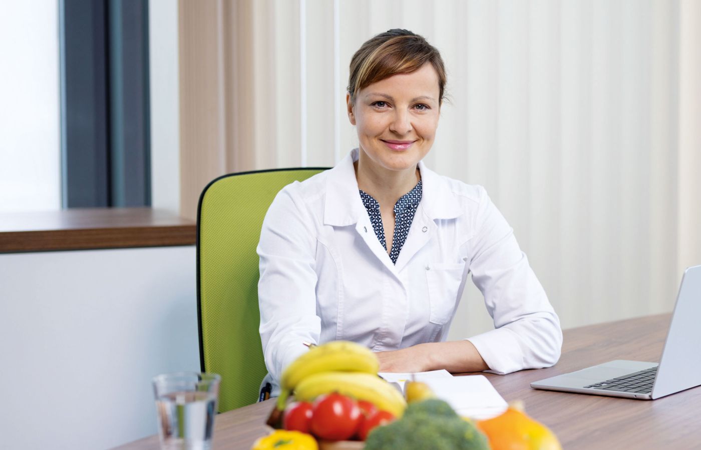 Ernährungsberaterin im weißen Kittel sitzt vor frischem Obst und Gemüse