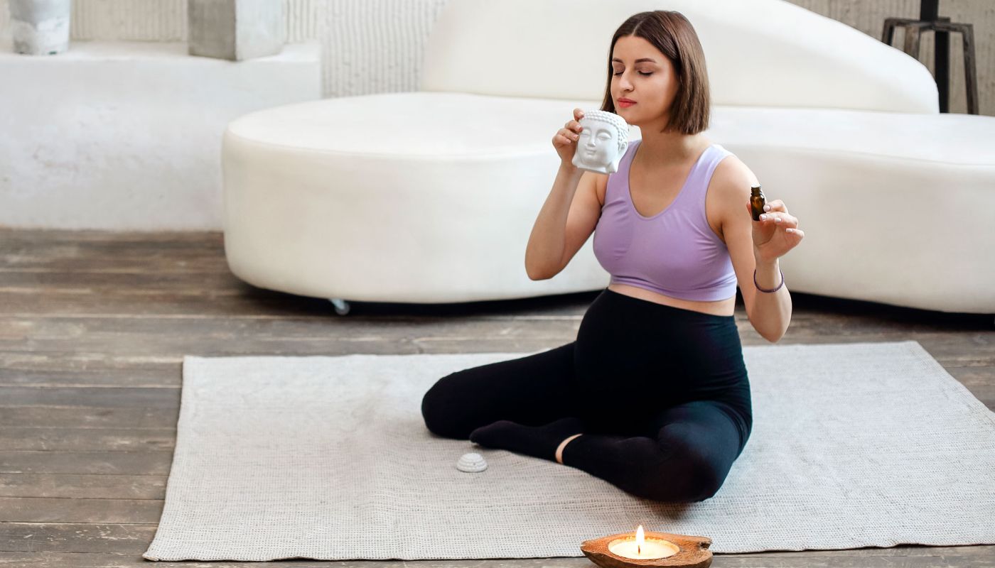 Schwangere Frau sitzt im Schneidersitz auf Yogamatte und riecht an einem Aromaöl