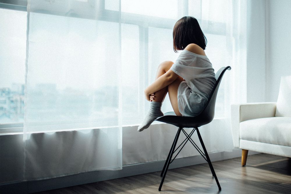 Frau sitzt allein auf einem Stuhl am Fenster, Kopf auf den Knien