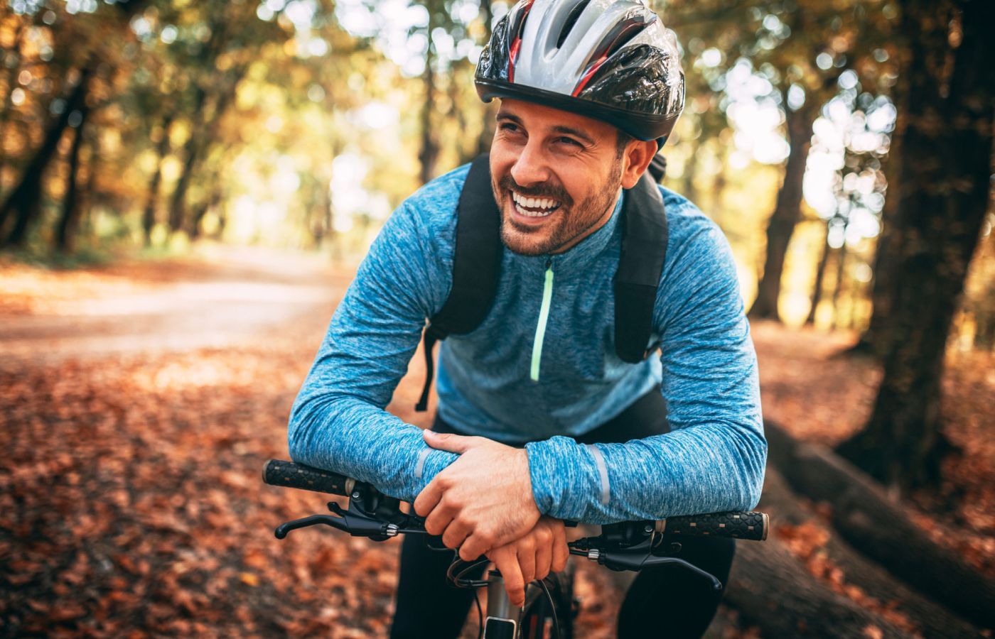 Lächelnder Mann mit Fahrradhelm und blauem Sportpullover lehnt sich auf sein Fahrrad, sonniger Waldweg im Hintergrund mit herbstlich gefärbten Bäumen.