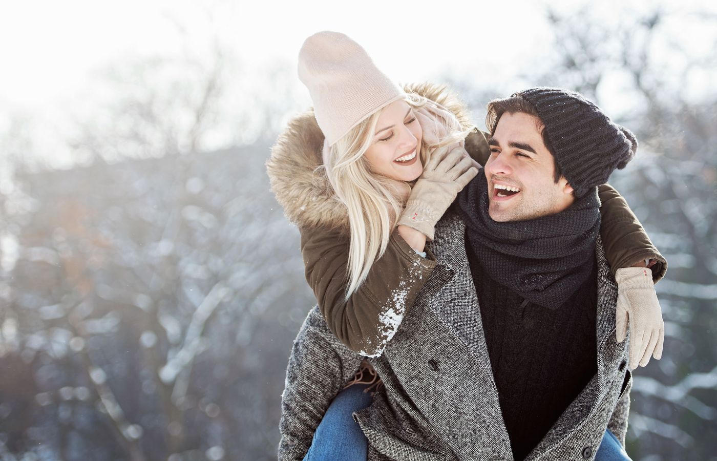 Zwei Personen in Winterkleidung lachen draußen im Schnee, eine Person trägt die andere huckepack.