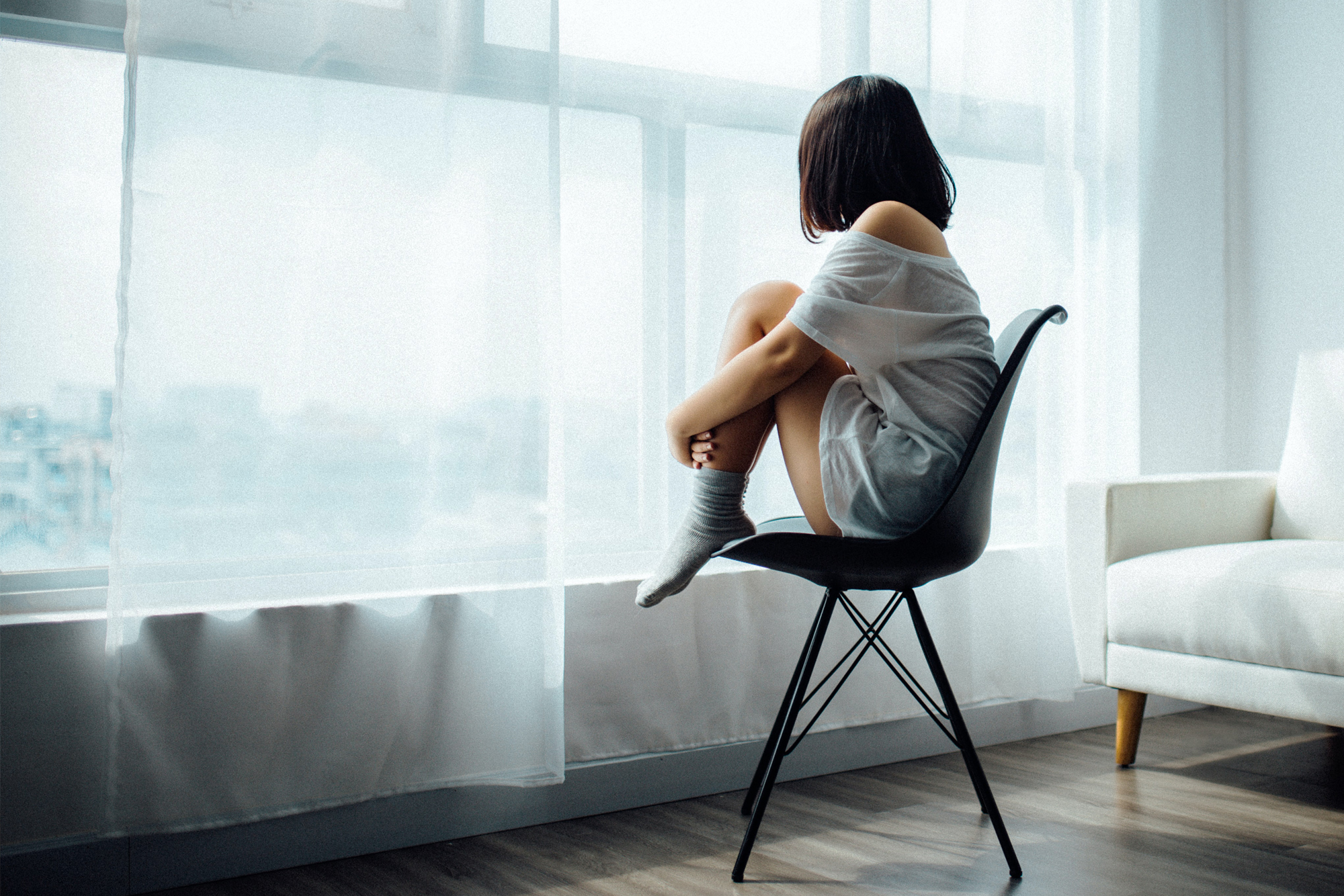 Frau sitzt allein auf einem Stuhl am Fenster, Kopf auf den Knien